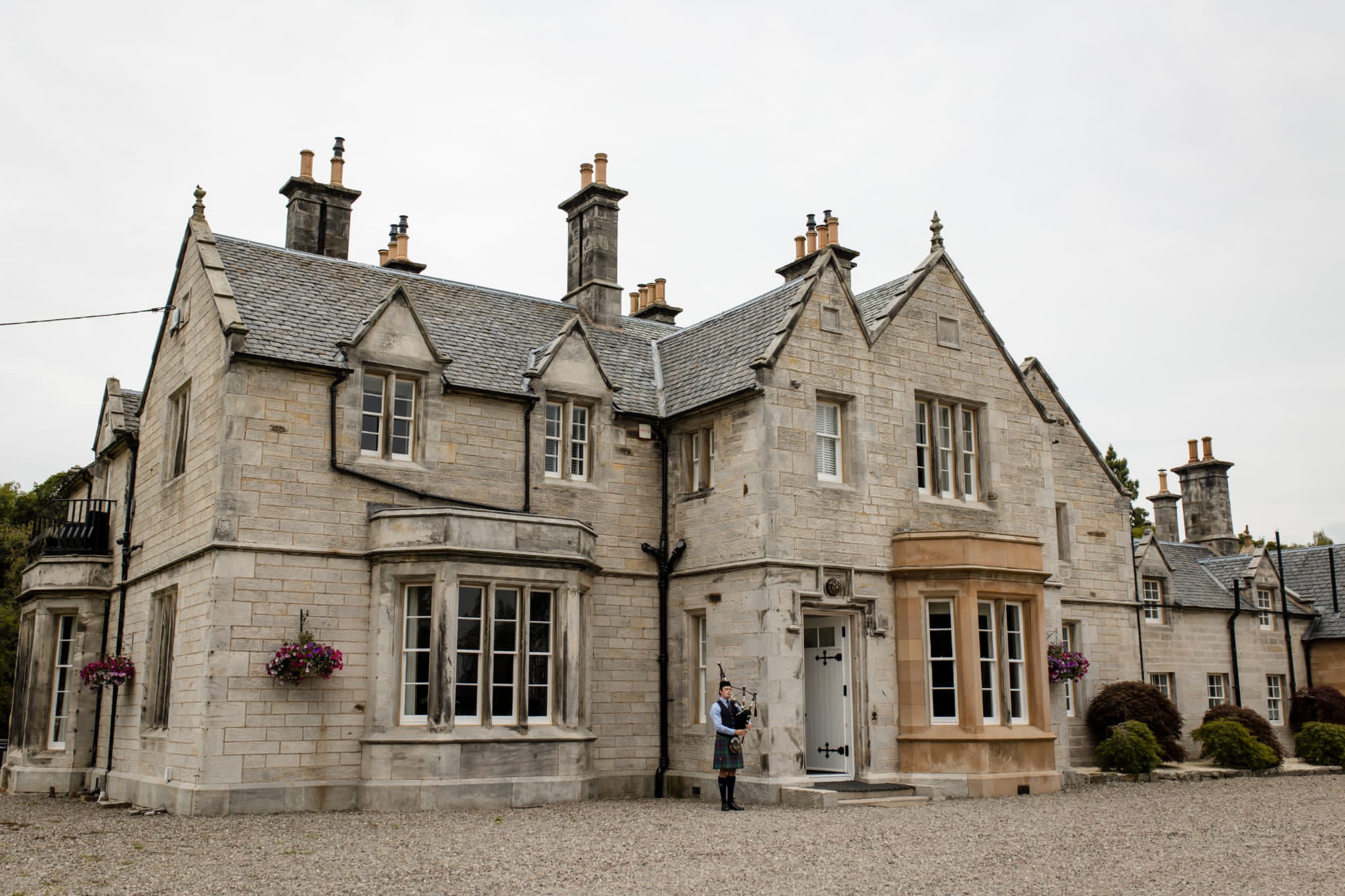 A Scottish Piper at a wedding piping guests into the ceremony. Wearing Traditional dress in from of a two story stone castle-like building with a red carpet entrance and a gravel driveway.