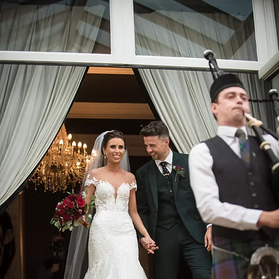 Scottish Piper Hire playing bagpipes whilst marching down stairs in front of a bride and groom inside a reception venue.
